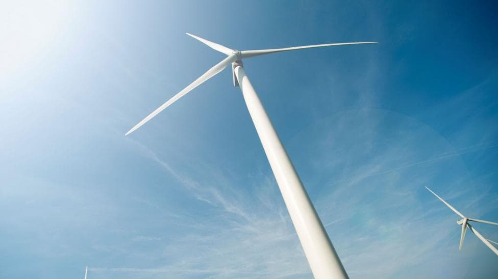 A view up a white wind turbine tower to its hub and long blades. There is another turbine nearby. The sky above is blue with wispy white cloud.