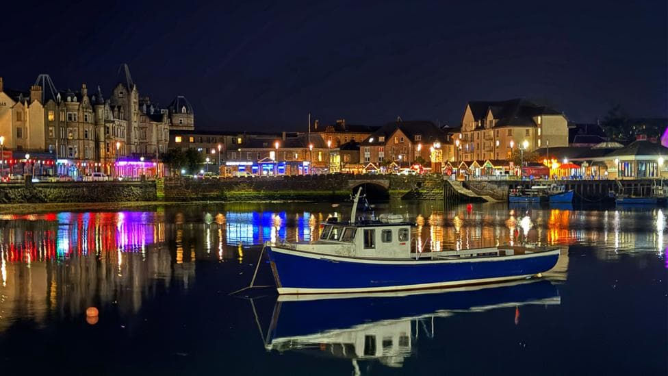 A blue fishing boat sits on calm water in Oban Harbour at night, with colourful lights from waterfront buildings reflecting across the water beneath a dark sky.