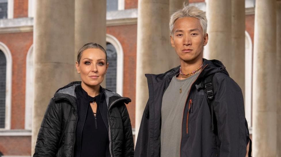 Carlos Gu and Amy Dowden pictured side by side in a promotional photo for the Channel 4 show, Celebrity Hunted. Carlos is wearing a grey dark coat, a light grey t-shirt and several necklaces. Amy is wearing a black top, jacket and coat. They are both looking at the camera and are not smiling.