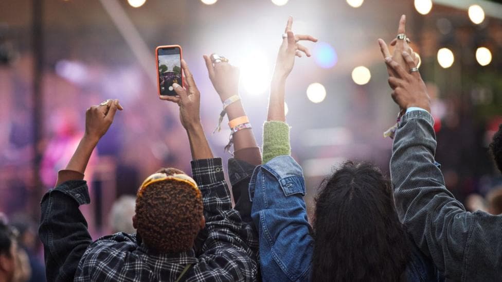 A group of young people have their backs turned to the camera and have their hands in the air as they attend a festival. One person is holding their phone up with the camera app open.