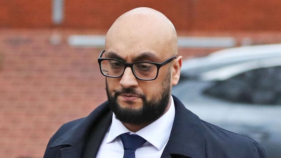 A man with spectacles and a dark beard walks across a street. He is wearing a jacket, white shirt and tie.