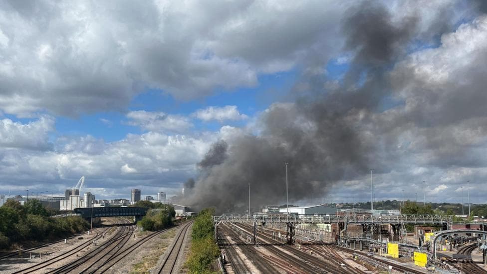 A thick plume of dark grey smoke rises into the sky above railway tracks, with Wembley Stadium's arch visible in the background.