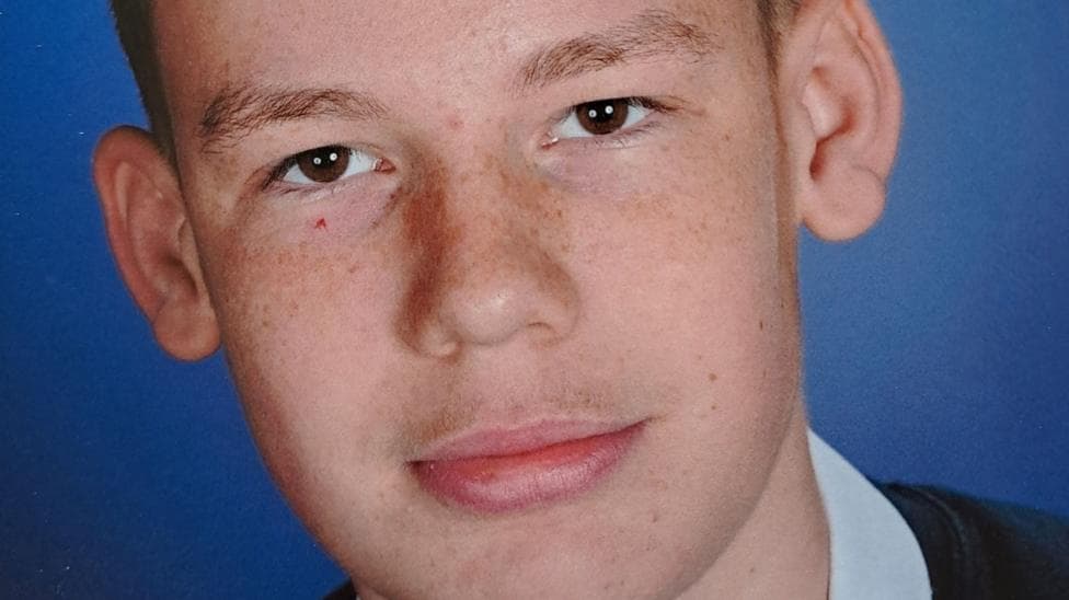 Close-up of Kenzie Carmichael who has short, dark hair. He is wearing a dark sweater over a light-collared shirt and tie