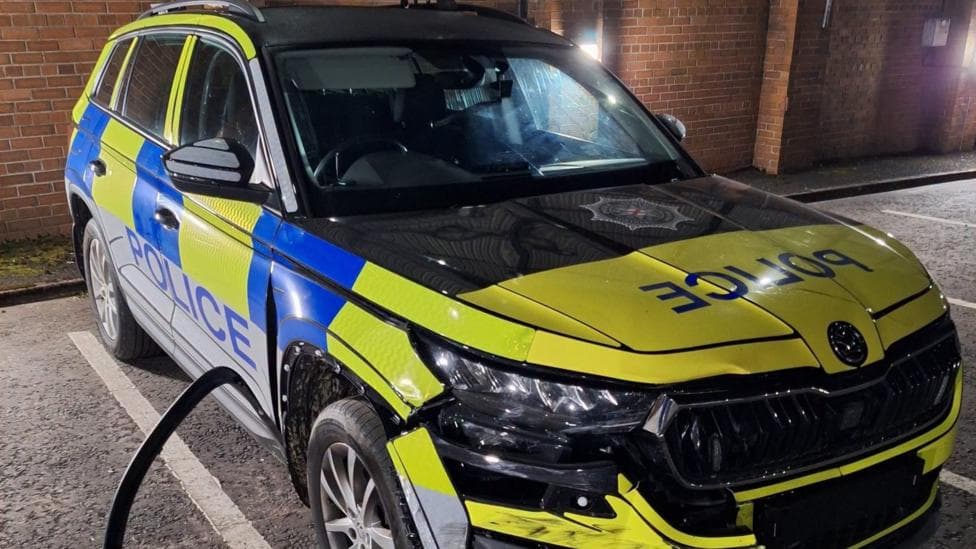 A yellow and blue police car pictured from the front, with significant damage to the bumper and side panel.