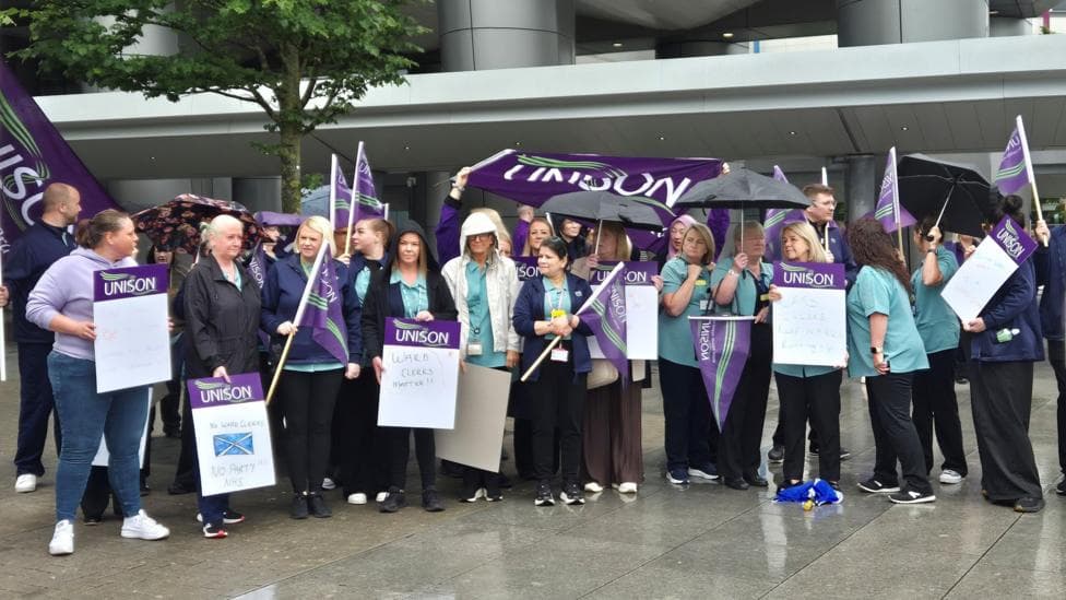 A group of hospital workers, holding signs and banners, outside a large hospital building