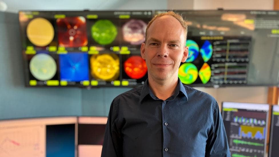 Richard wears a dark blue shirt which is unbuttoned at the collar. He is looking towards the camera with several screens showing images of the sun behind him