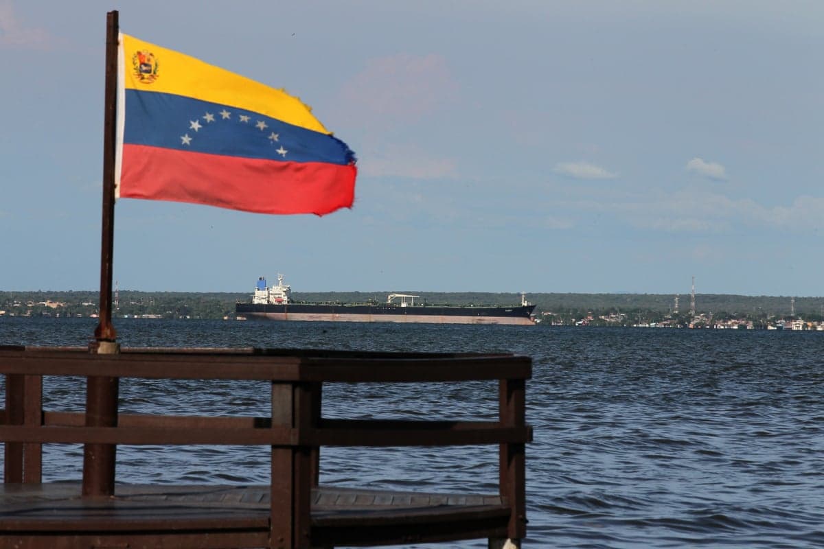 Red, yellow, and blue flag in wooden pier, with choppy water and very long ship beyond.
