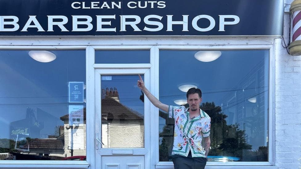 A man in a white floral shirt standing in front of a barber shop. He is pointing at the sign.