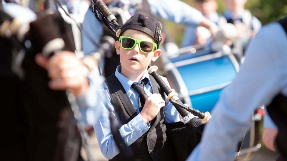 A young musician playing pipes and taking part in a parade. He is wearing neon green sunglasses, a black waistcoat, hat and tie