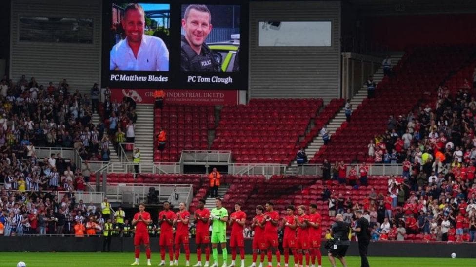 Rear view of a football team - wearing red strips - standing to attention in front of a bank of stadium seats, with other players to the side. A screen above shows the image of two men.
