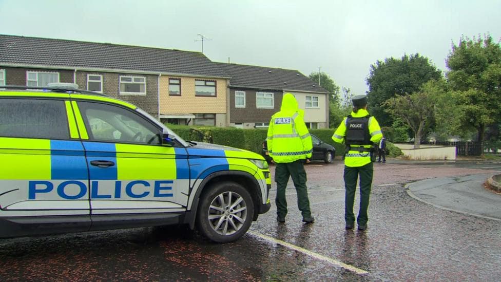 Two police officers stand beside a car, wearing hi-vis waterproof coats and police vests.