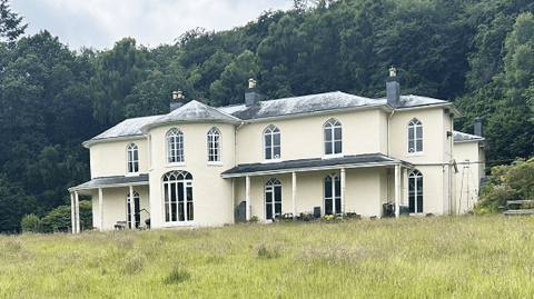Dol-llys Hall taken from the far side of a long-grass lawn. The hall is painted light yellowy cream and has a slate tiled roof. To the left and right of a round tower-like protuberance are two covered verandas with supporting posts. Six large arched white-framed leaded windows are visible on both floors, with three chimney sets on the roof. The house is framed by large, mature woods. Some washing is visible on a line to the right of the picture.