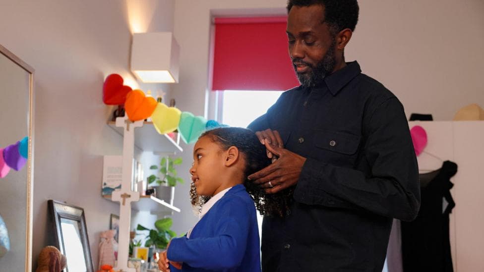 A man wearing a black shirt tying his daughter's hair up, she is wearing a blue school uniform with a white shirt.