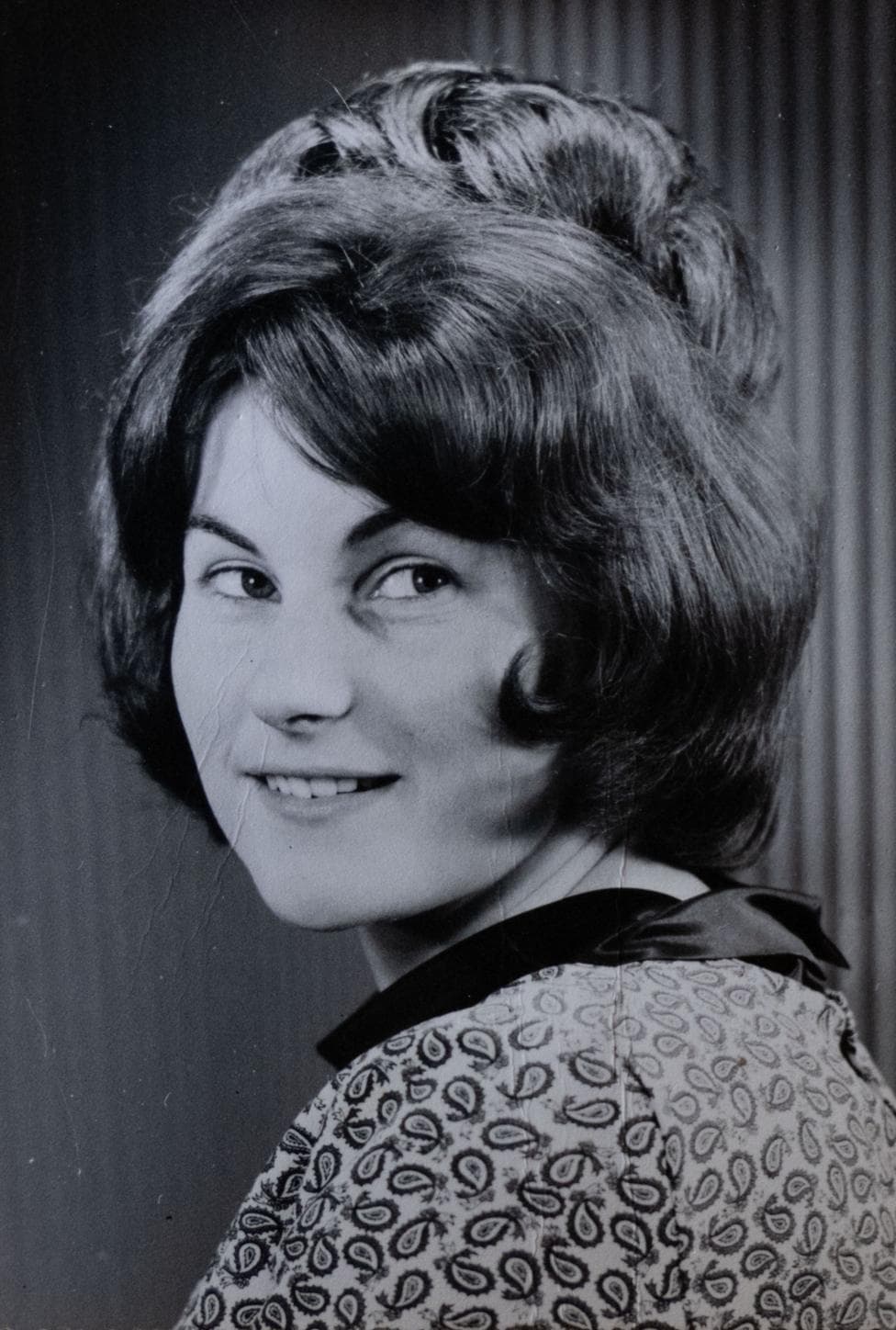 A posed black-and-white image of a young woman, looking over her shoulder. She has a lot of dark hair and is wearing a paisley-patterned top.