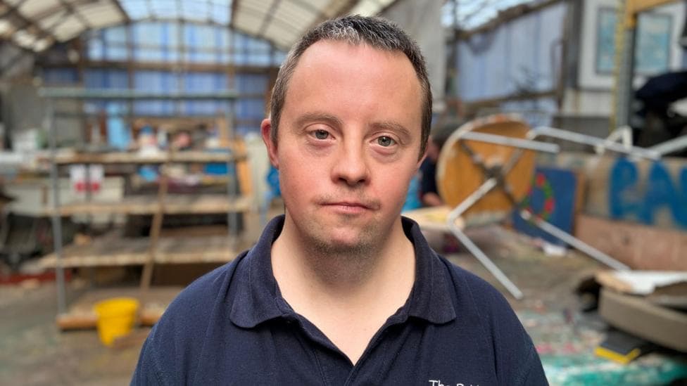 A man with short dark hair stands in a bright, spacious workshop filled with creative energy. He wears a navy-blue collared shirt embroidered with “The Bridge Association” and looks directly at the camera. Behind him, shelves of materials, colourful artwork, and scattered tools hint at an active, hands-on environment.