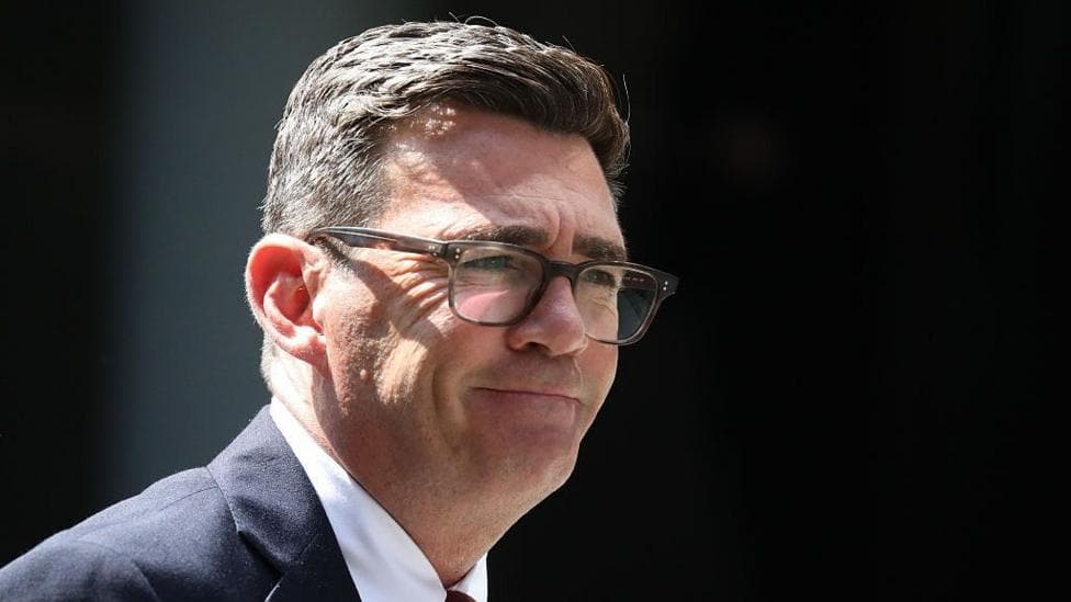 Andy Burnham wearing brown glasses and a navy suit standing outside in the sun, smiling with his mouth closed.