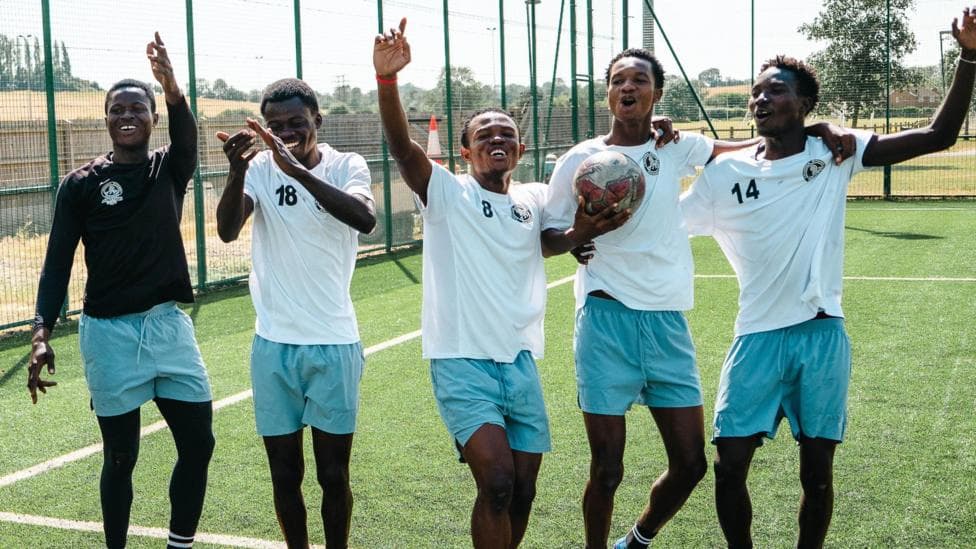 Five young men stand in a line looking towards the camera, with four wearing white T-shirts and one in a black t-shirt and all in turquoise shorts, smile and hold their hands up as they are captured midway through singing a song. One holds a football while they stand on an artificial football pitch with fencing visible around it in the background