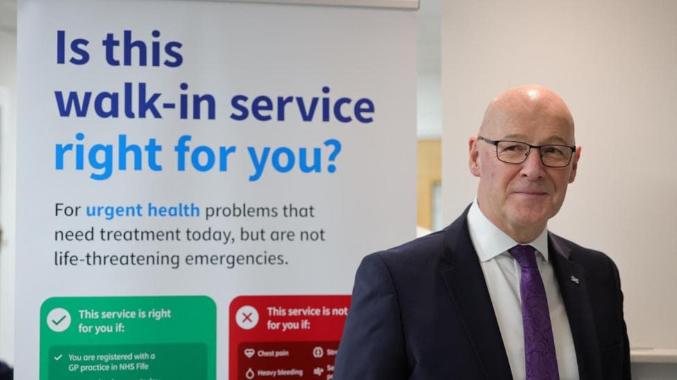 First Minister John Swinney during a visit to officially open the new GP walk-in service at Randolph Wemyss Memorial Hospital in Leven, Fife - A person in a suit and tie stands beside an NHS Fife information board promoting a GP walk-in service for urgent, non-life-threatening health problems. The poster explains who can use the service and outlines suitable and unsuitable conditions for treatment.