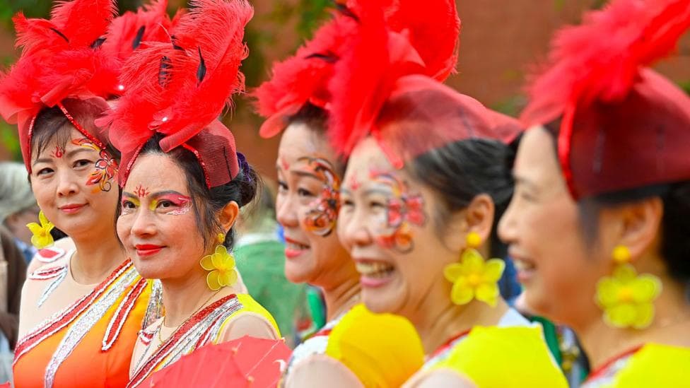 Five women are wearing bright yellow and orange clothing. They have red hats on with feathers.
