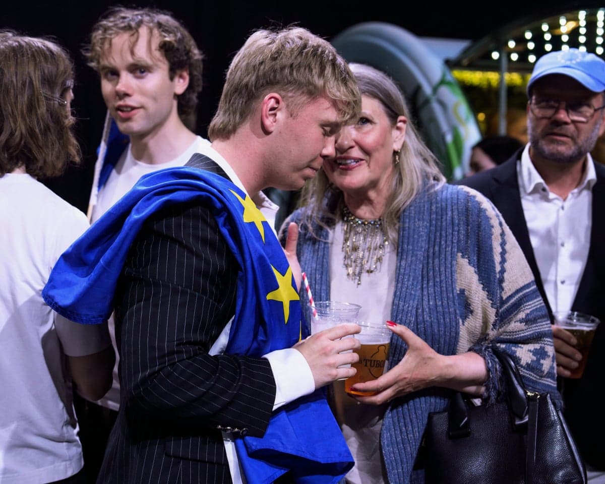 A man with an EU flag over his shoulder speaks to a woman at a drinks party.