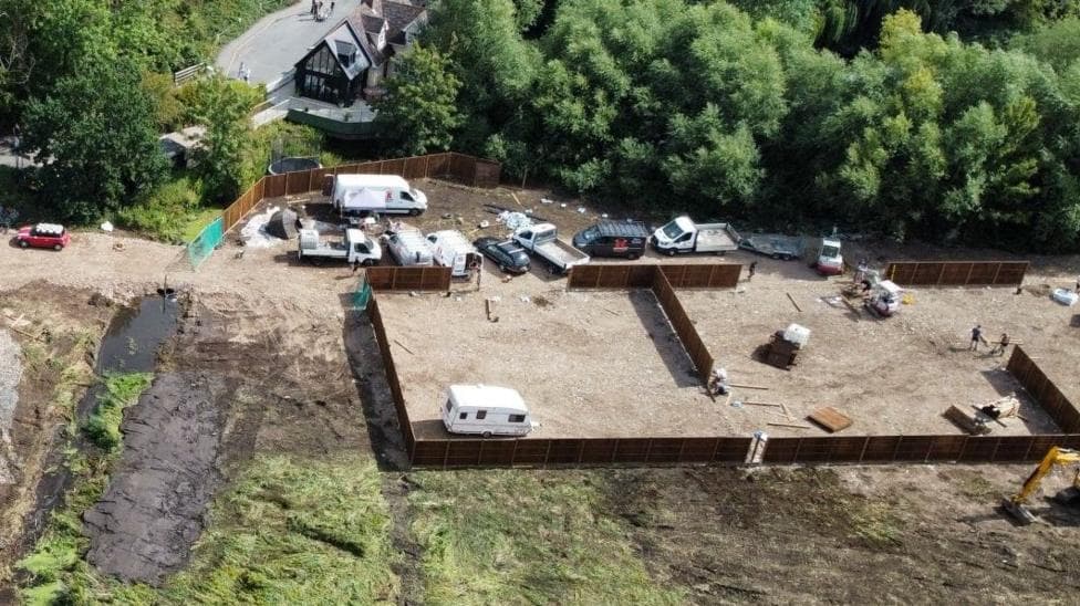 A drone picture of the field, which has been badly damaged and covered with hardcore. There are lots of vans on site and a caravan. It is surrounded by brown fencing.