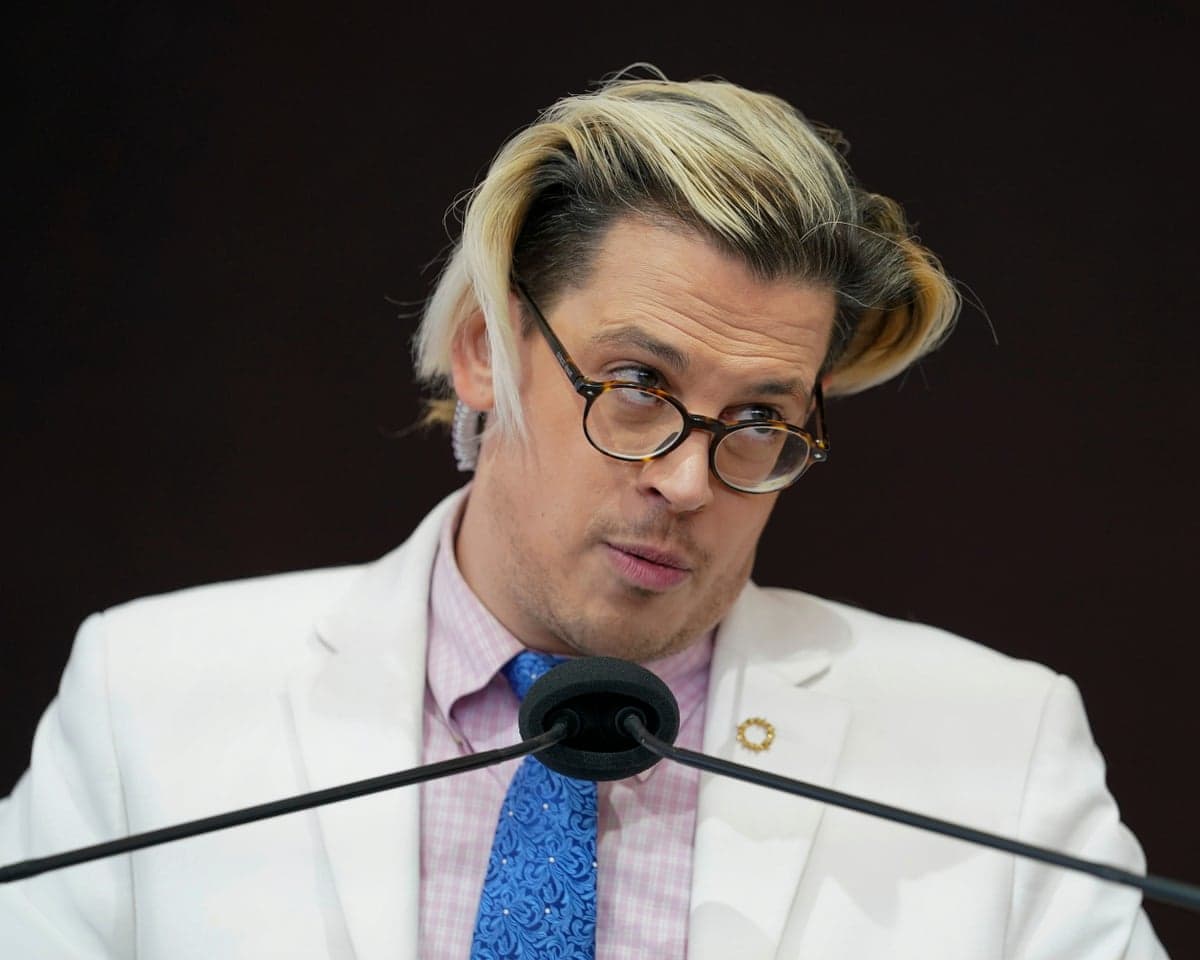 Yiannopoulos speaks into a microphone at a podium, wearing a white blazer and blue tie