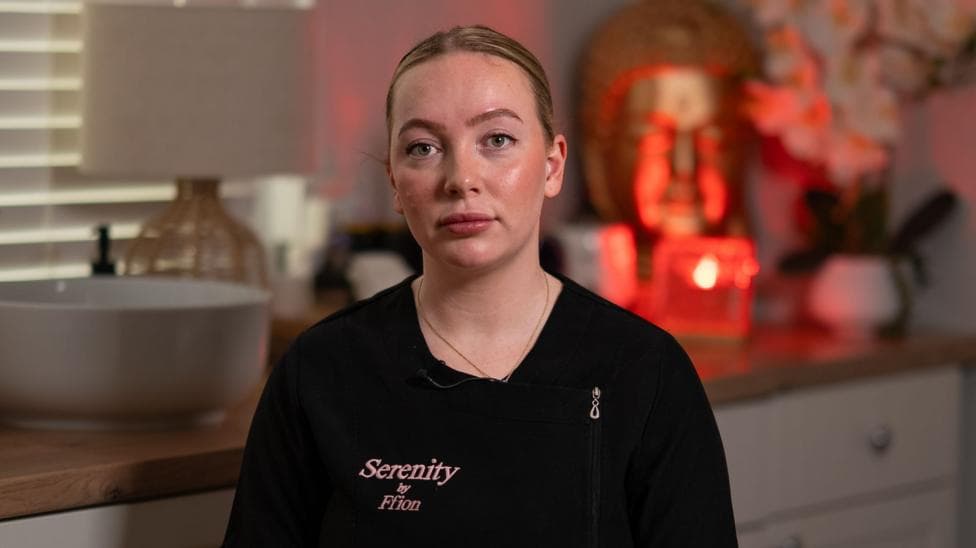 Ffion looks at the camera with a stern expression. She wears a black uniform with a pink Serenity by Ffion logo. She has her blonde hair in a slick back bun and wears a gold necklace. She is sat in front of a work place which has a sink on, alongside plants and a red mood light.
