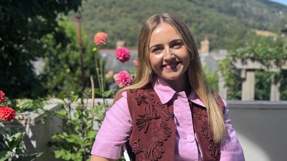 Hannah is a woman in her early 30s. She has long blonde hair and is smiling at the camera. She's pictured in front of flowering plants