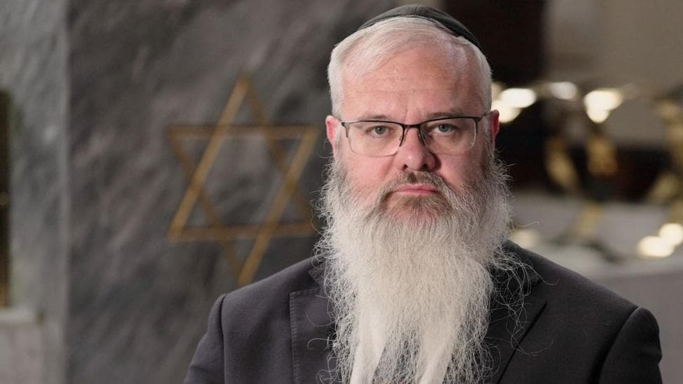 Rabbi Daniel Walker, who is wearing a black blazer, shirt and tie and a kippah on his head, is starring directly at the camera.