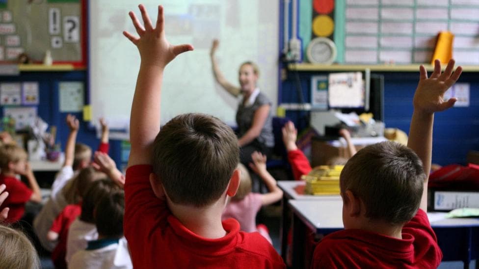 In a primary school classroom, two young boys in red tops with short hair stick their hands up in the air in the foreground, while their classmates who are clustered in front of them sitting down also stick their hands up in the air. An out-of-focus teacher with blonde hair and a grey top in front of a whiteboard also has her hands up and is smiling at the children in front of her. The classroom has blue walls and lots of posters on the walls and white school tables moved out of the way in the right hand of the image.