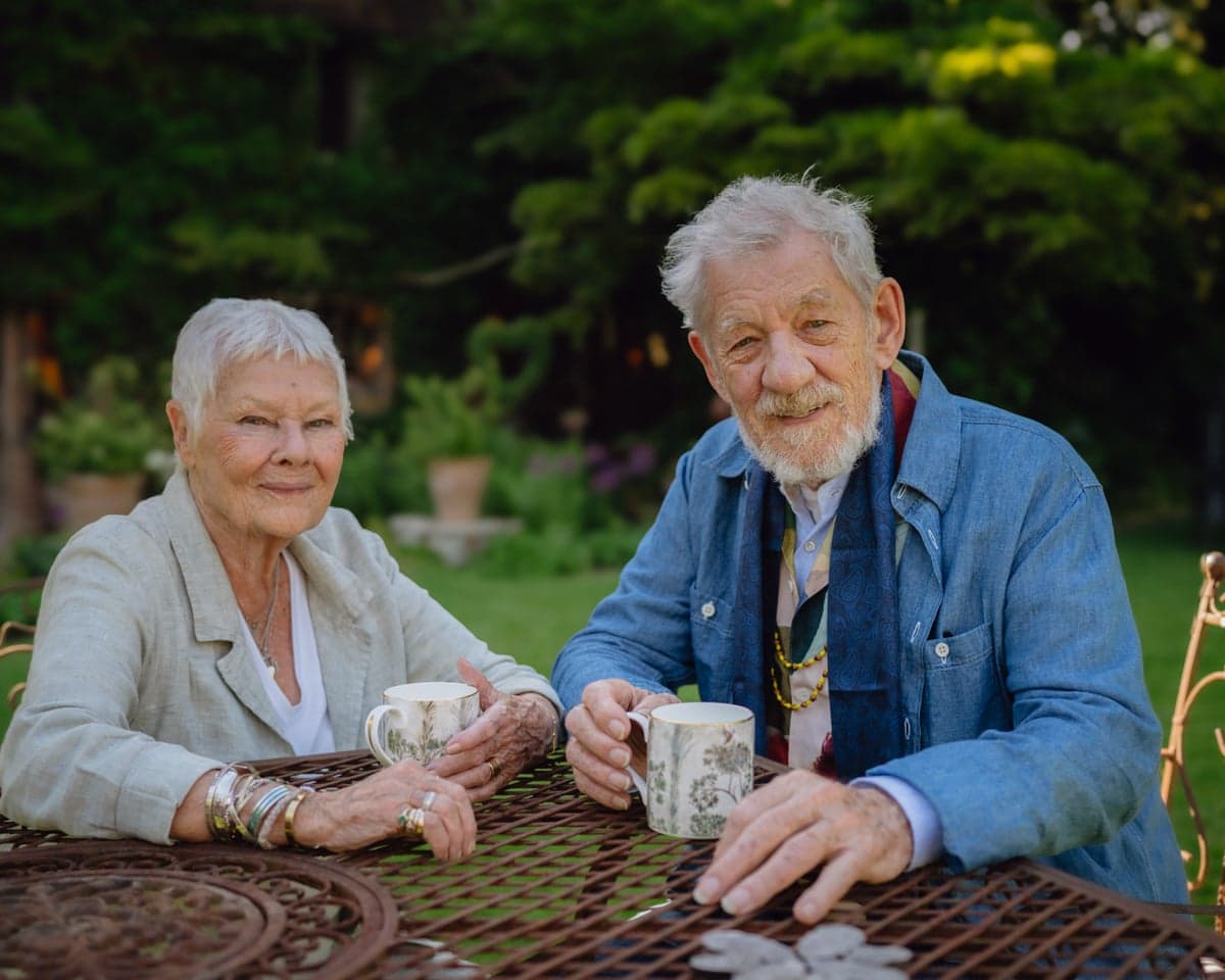Judi Dench and Ian McKellen smile at an outdoor table holding mugs, greenery behind them