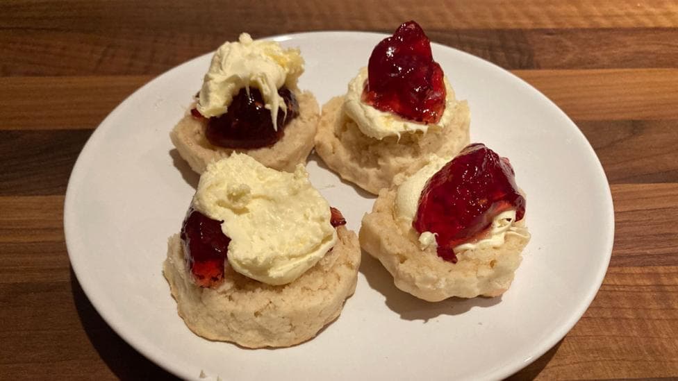 Two scones split in half with cream and jam applied on each half. Two of the halves have jam on first then cream on top and the other two have cream and jam the opposite way.
