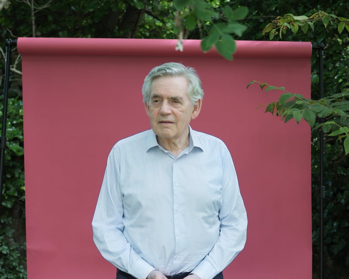 Gordon Brown, with white hair and a light blue shirt, stands before a pink outdoor backdrop