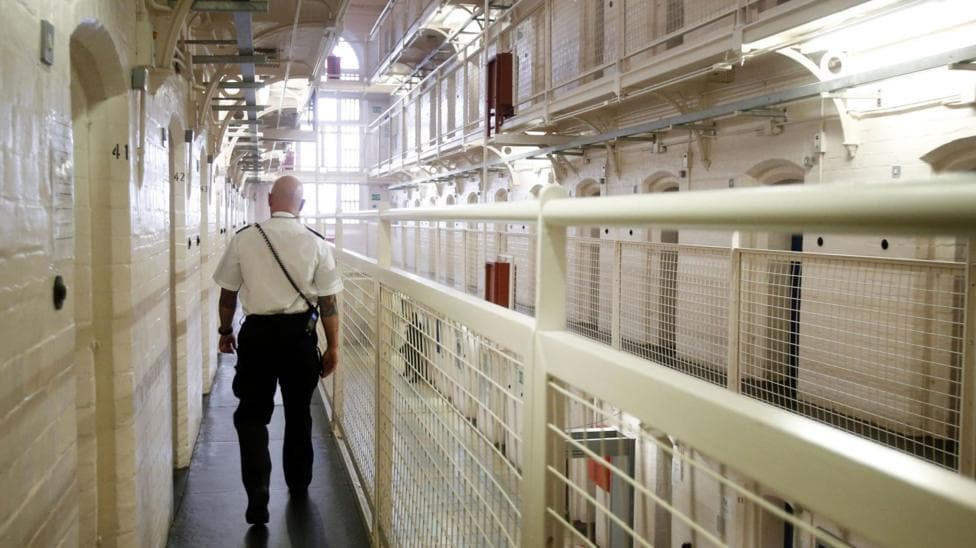 The interior of a prison, it has cream walls and metal fences. A prison guard walks up the corridor.