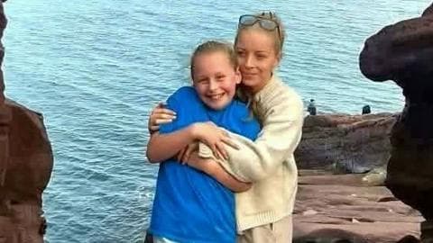A woman with tied-back blonde hair with sunglasses on her head hugs a young girl, who is wearing a blue t shirt and white shorts. The woman is wearing a cream cardigan and light shorts. They are framed by cliffs and the North Sea behind them