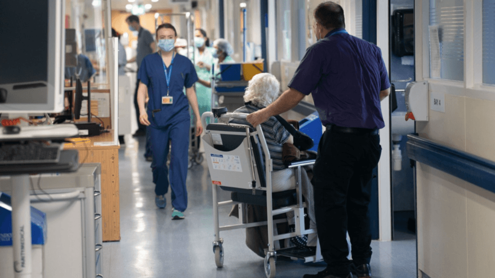 Hospital corridor with healthcare staff and equipment visible in the background, while a seated patient is being pushed in a wheeled hospital chair by another person. A staff member in blue scrubs and a face mask walks along the corridor, with treatment rooms and medical workstations lining both sides.