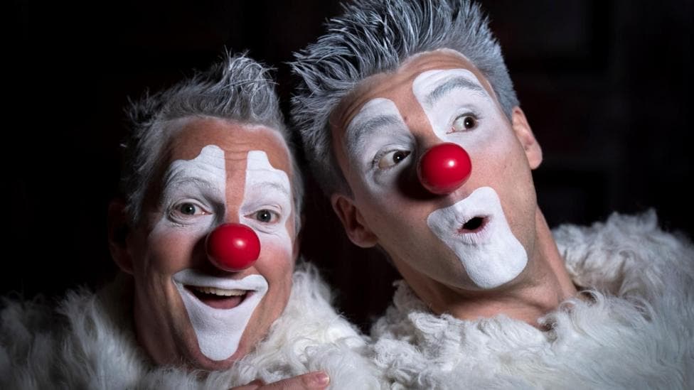 Remi Jacques and Jean-Felix Belanger with their faces painted as clowns. They have exaggerated white make-up around their mouth and eyes and are wearing red, round fake noses. Their hair is spiked up and sprayed silver and they are wearing fluffy, white outfits.