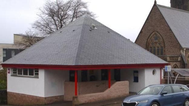 The picture shows Fort William's Viewforth public toilet block. It has white walls and bright red supports by the entrance. A church building is visible in the backgrounds.