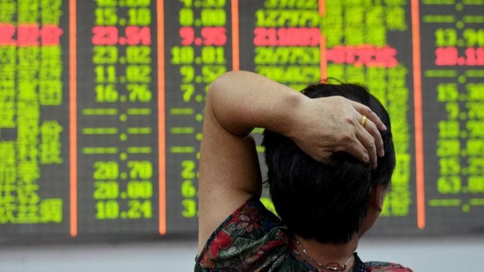 An investor looks at an electronic board showing stock information with numbers highlighted in red and lime green against a black background. The investor has her back to the camera and has her left hand resting on her head.