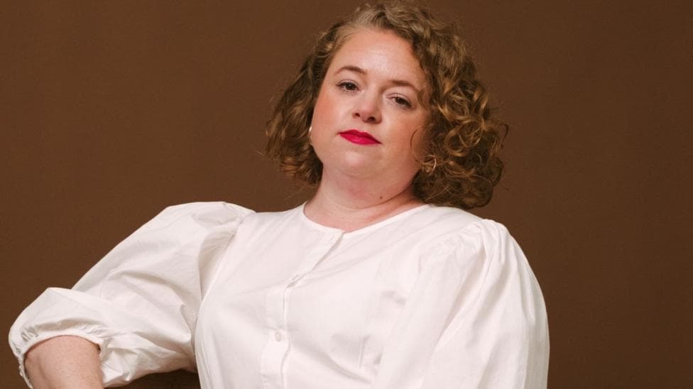 Woman with curly red hair and bright red lipstick faces the camera. She is wearing a white shirt