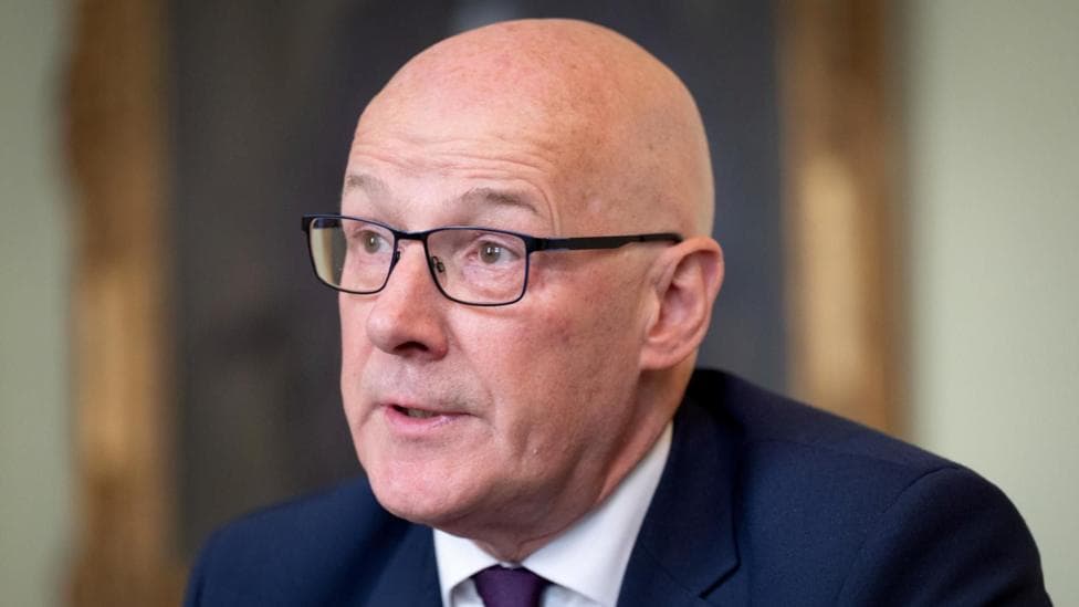 John Swinney has a bald head and is wearing black-rimmed glasses. He is wearing a blue suit jacket, white shirt and purple tie. He is looking to the side of the camera