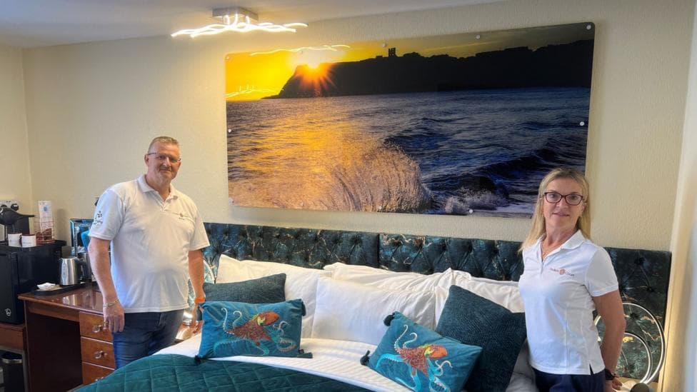 This picture shows James and Angela Rusden - who run a B&B once voted the world's best - preparing one of the rooms. James, on the left of the image, has short brown hair, is wearing a white polo-neck T-shirt and a gold bracelet. Angela, on the right, has shoulder-length blonde hair and is wearing a gold necklace and a white polo-neck T-shirt. A huge photo of Scarborough Castle and the town's North Bay is hanging on the wall.