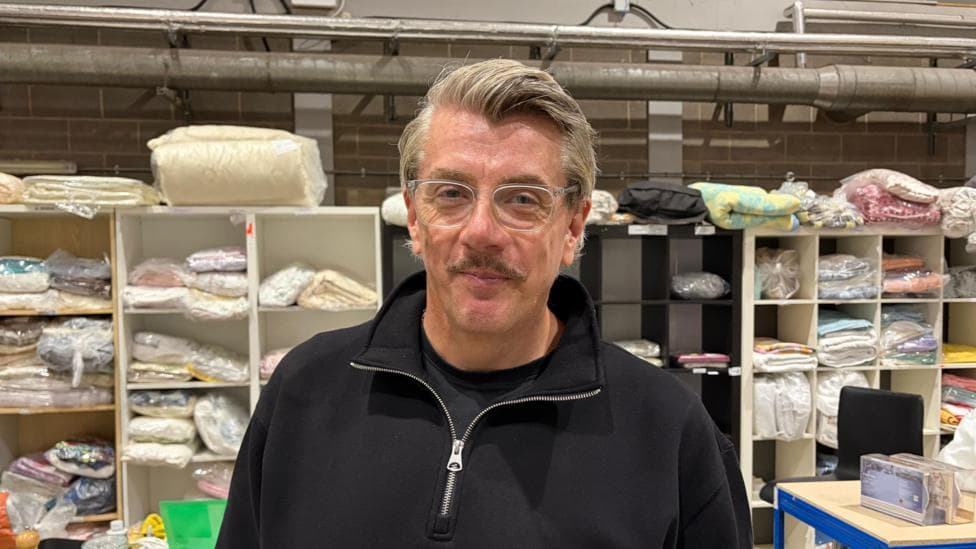 Ashley is looking at the camera with a neutral expression, in the background are shelves with sheets wrapped in plastic.