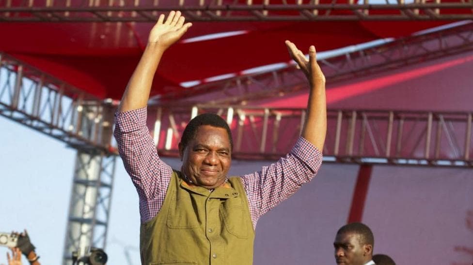 Hakainde Hichilema with an olive waistcoat over a checked shirt holds his arms aloft.