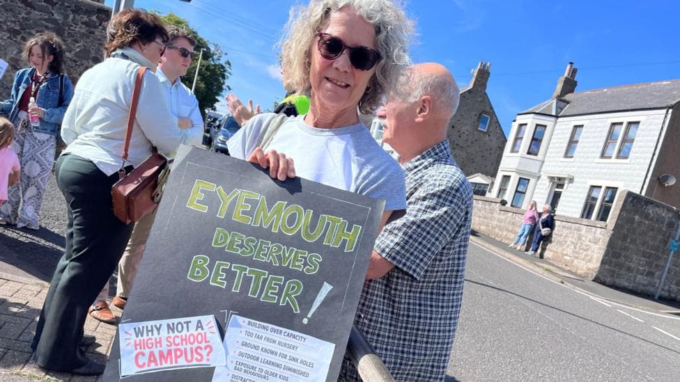 A grey-haired woman holds a sign saying 'Eyemouth deserves better' with people in the background