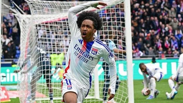 Malick Fofana, wearing Lyon's white kit with a red-and-blue stripe, celebrates scoring a goal by waving his right arm in the air