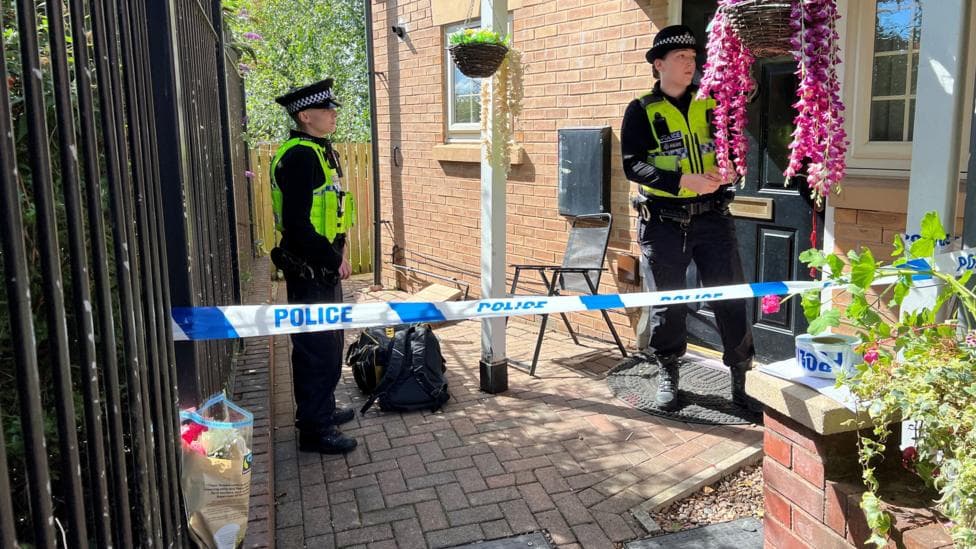 Two police officers stand guard outside the entrance to a house. Visible in the photo is some police tape and a bouquet of flowers.