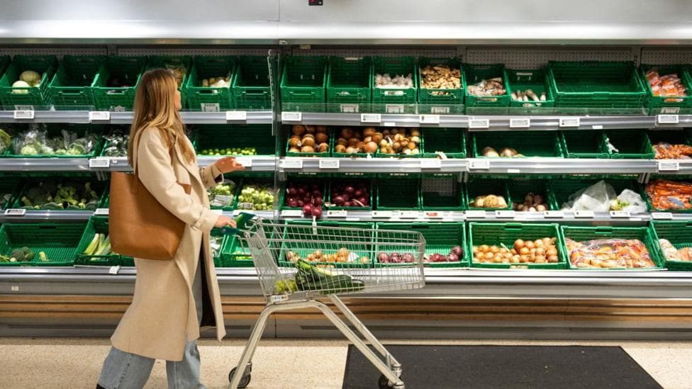 A woman pushing a trolley past the vegetable section of a supermarket