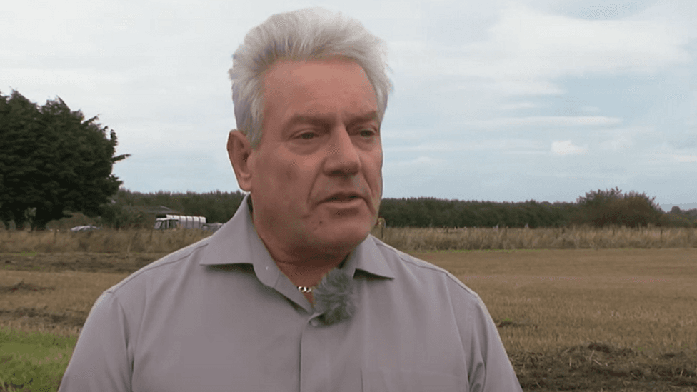 A man with short grey hair and a light brown shirt is wearing a microphone while standing next to a field. He is being interviewed. Two vehicles can be seen on a road behind him.