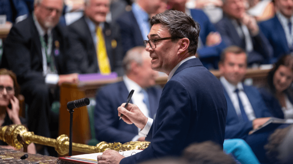 Andy Burnham speaking in the House of Commons. He is wearing a dark suit and holding a pen.